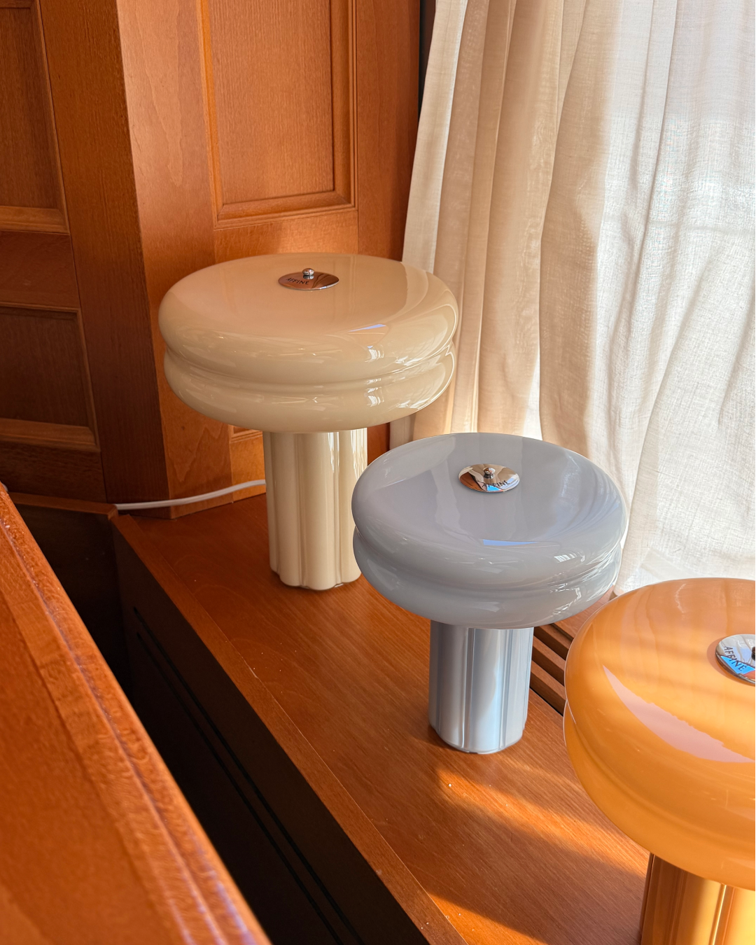 The Affiné Table Lamp in Crème Beige styled together with the Portable Lamp in Soft Amber and Powder Blue beside a wooden-framed window.