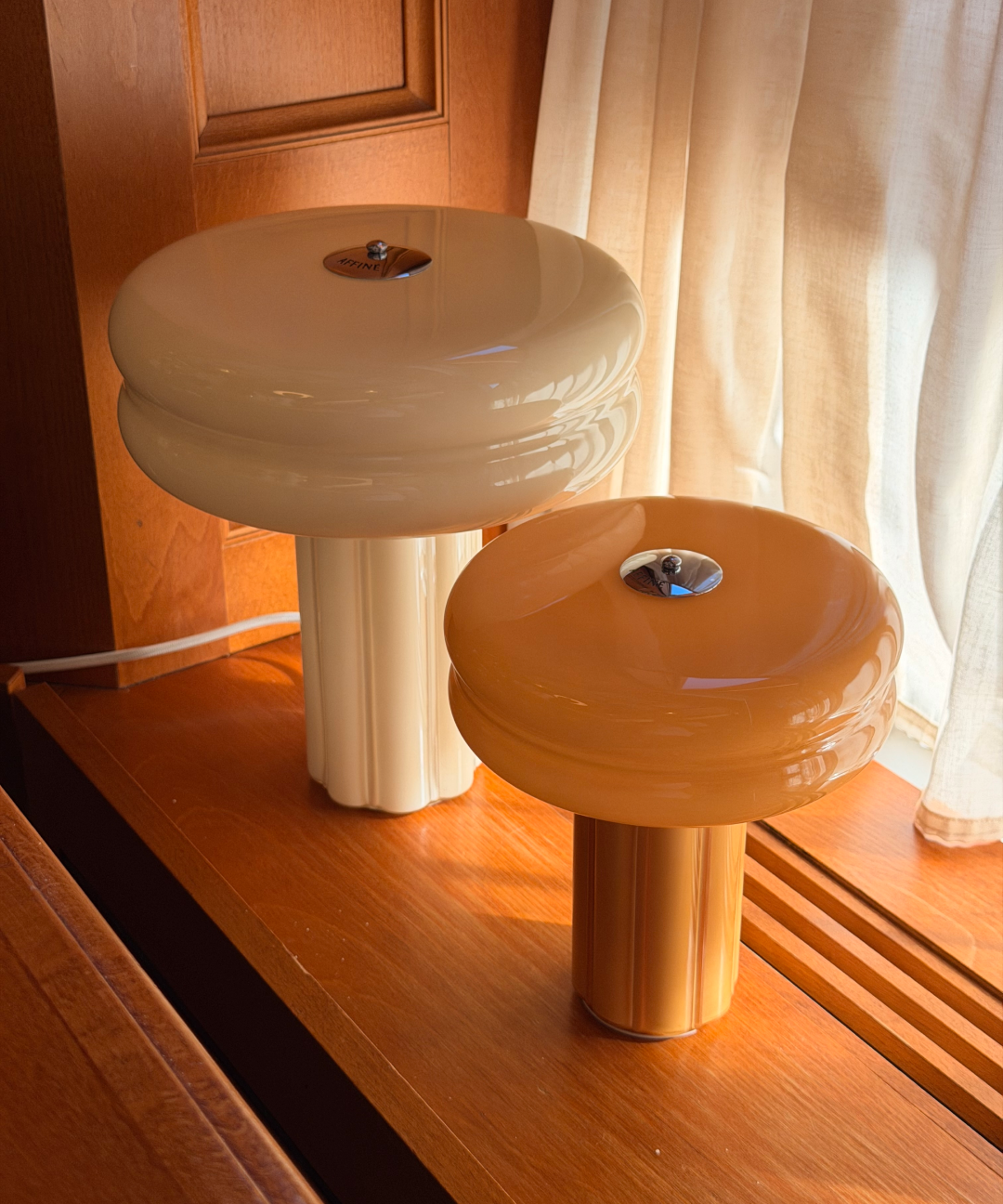 The Affiné Table Lamp in Crème Beige and Portable Lamp in Soft Amber styled together by a wooden-framed window.