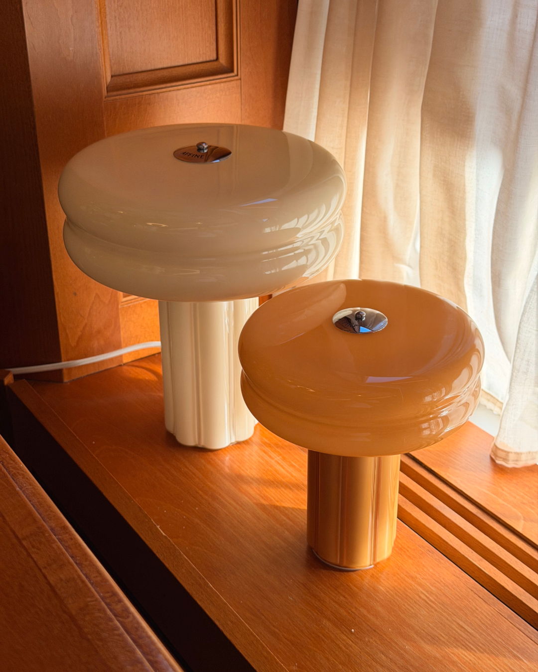 The Affiné Table Lamp in Crème Beige and Portable Lamp in Soft Amber styled together by a wooden-framed window.